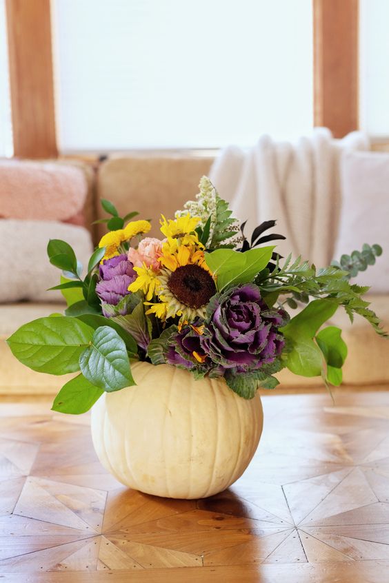 A White Pumpkin With Foliage, Faux Yellow And Purple Blooms And Some Dried Touches Is A Chic Fall Centerpiece