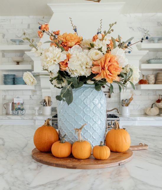 An Elegant White And Orange Faux Flower Arrangement In A Blue Vase And Orange Pumpkins Around It