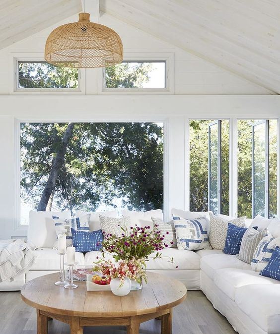 a beuatiful modern farmhouse coastal sunroom with a large sectional, blue pillows, a wooden table and woven chandelier