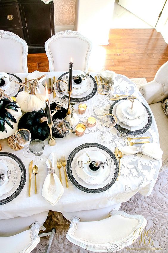 A Glam Halloween Tablescape With White And Black Pumpkins, Black Feathers, Candles, Gold Cutlery And Black Chargers