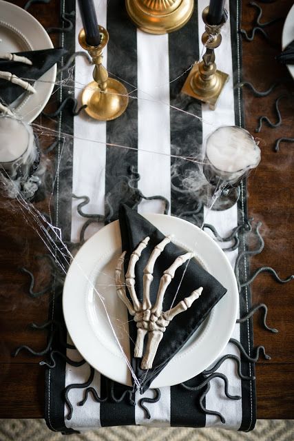 A Black And White Halloween Table With A Striped Table Runner, Black Napkins, Skeleton Hands, Gold Candleholders And Snakes