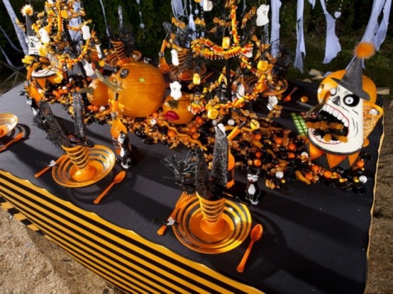 An Extra Bright Blakc, Graphite Grey And Orange Halloween Tablescape With Orange Plates, Cutlery, Scary Pumpkins And Faces, Lots Of Candies And Witches' Hats