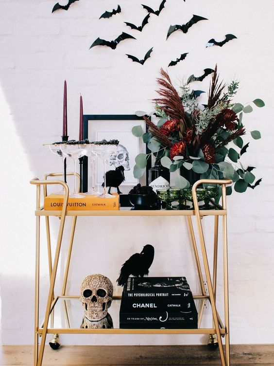 A Halloween Bar Cart With An Embellished Skull, A Dark Floral Arrangement, Burgundy Candles And Bats Over The Cart