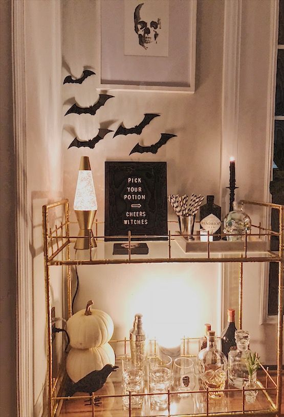 An Elegant Halloween Bar Cart With Bats On The Wall, Lamps And Candles, White Pumpkins, A Sign And A Crow