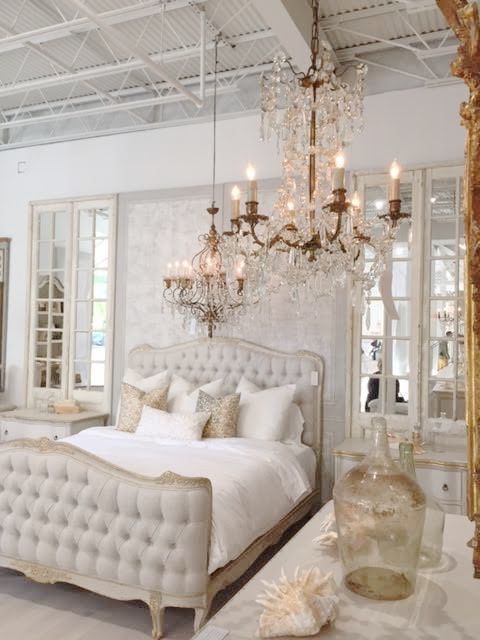 A Refined Neutral Bedroom With A Chic Bed, Crystal Chandeliers, Mirrors And Printed Pillows