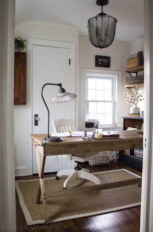 A Rustic Vintage Home Office With A Wooden Trestle Desk, Vintage Lamps, A White Chair And Cotton Branches In A Vase