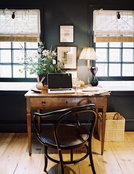 A Vintage Rustic Home Office With Black Walls, Woven Shutters, A Wooden Desk, A Black Chair, Lamps And Greenery