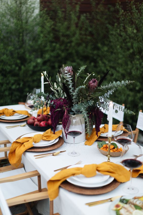 A Bright Modern Thanksgiving Table With Greenery And Deep Purple Blooms, Yellow Napkins, Woven Chargers And Gold Cutlery Rocks