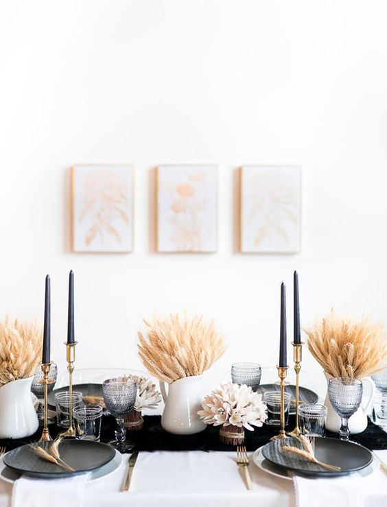 A Modern Thanksgiving Tablescape With A Black Runner, Plates And Tall Candles, Wheat Arrangements And Grey Glasses