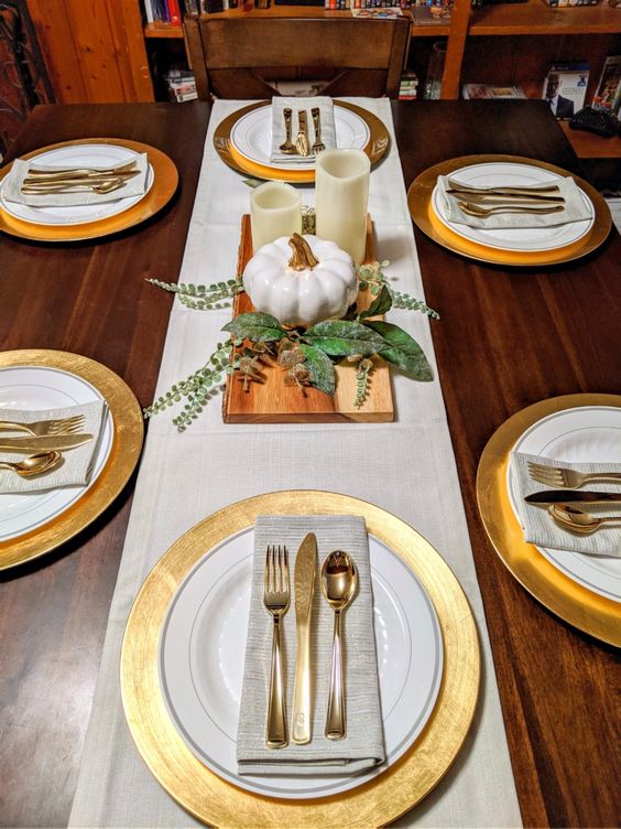 A Modern Rustic Thanksgiving Tablescape With Gold Chargers, Neutral Linens, White Candles And Pumpkins And Some Greenery