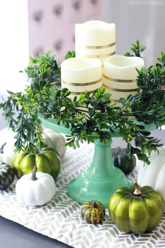 A Modern Thanksgivving Centerpiece Of A Green Stand With Greenery, Striped Candles, White And Green Pumpkins And A Printed Runner