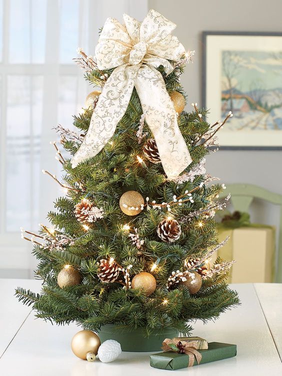 A Tabletop Christmas Tree Decorated With Lights, Willow, Snowy Pinecones, A Beautiful Patterned Bow On Top And Gold Ornaments