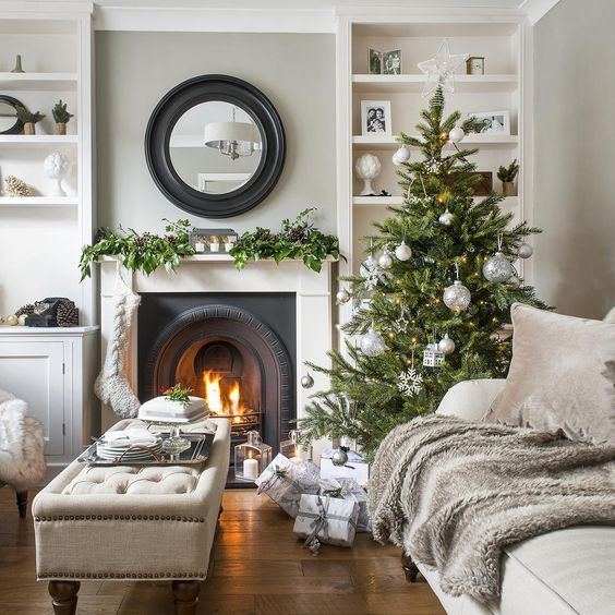 A Stylish Christmas Tree With Silver Ornaments, A Stocking And A Greeneyr Garland With Pinecones On The Mantel